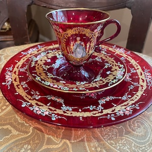 Puede incluir: Juego de taza y platillo rojo rubí con ribetes dorados y diseños florales y foliares ornamentados. La taza presenta una escena pintada de una mujer con un vestido. El juego se exhibe sobre una superficie dorada estampada.