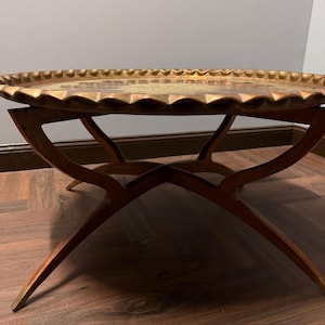 May include: A round coffee table featuring a large, circular brass tray top with a scalloped, wavy edge. The tray rests on a wooden base composed of four curved, intersecting legs that form an X-shape, providing a stable support structure.