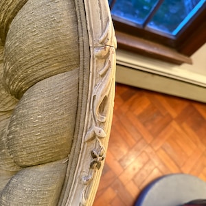 May include: Close-up of a section of an antique armchair. The chair features a light green, textured fabric and a cream-colored, ornate wooden frame with carved floral details. The frame shows signs of wear, adding to its vintage appeal.