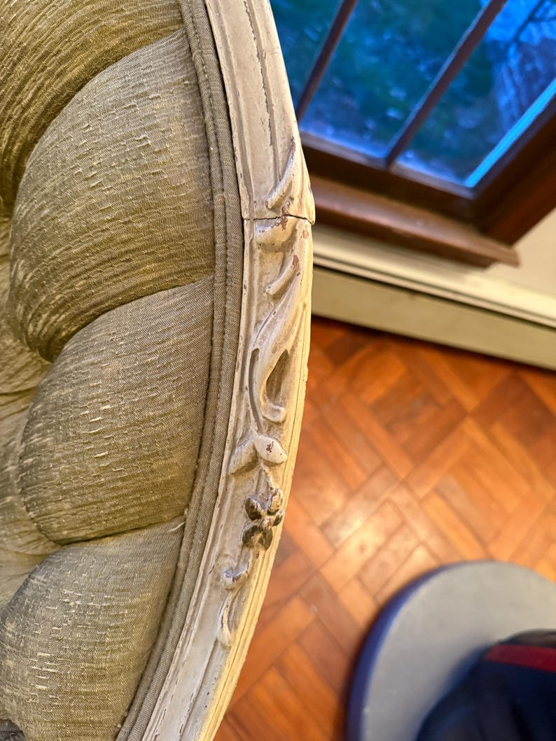 May include: Close-up of a section of an antique armchair. The chair features a light green, textured fabric and a cream-colored, ornate wooden frame with carved floral details. The frame shows signs of wear, adding to its vintage appeal.