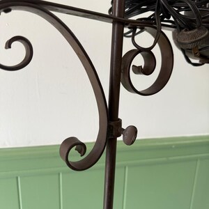 May include: Close-up of a dark brown metal floor lamp with decorative scrollwork. The lamp features a central pole with a curved arm and a small knob. A black electrical cord is visible.
