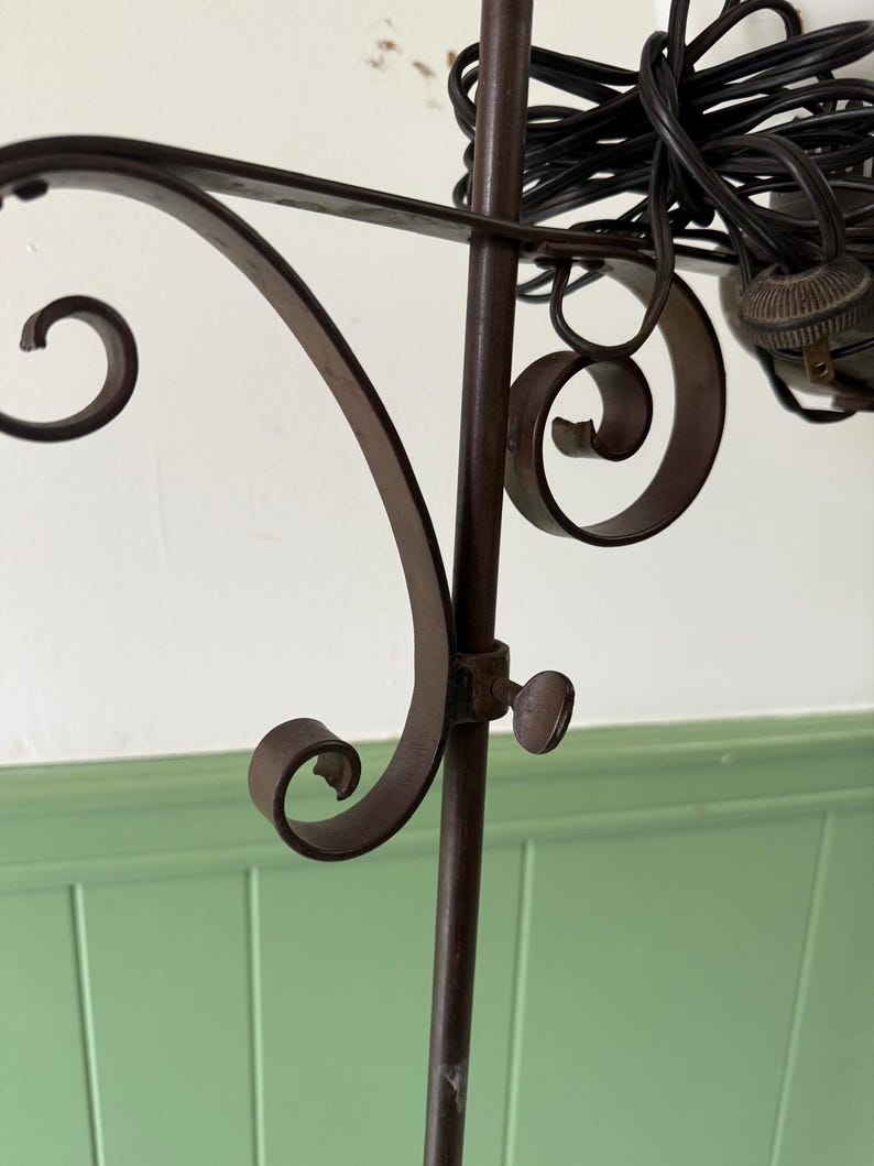 May include: Close-up of a dark brown metal floor lamp with decorative scrollwork. The lamp features a central pole with a curved arm and a small knob. A black electrical cord is visible.