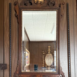 May include: A rectangular, ornate wooden mirror with a gold shell-shaped accent at the top. The frame is dark brown with intricate carvings. The mirror is mounted on a wood-paneled wall.