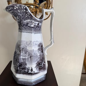 Peut inclure: Une cruche en céramique blanche et grise avec un motif paysager détaillé. La cruche comporte une anse et un bec verseur, et le motif comprend des scènes de bâtiments, d'arbres et de personnes. La cruche est posée sur une surface en bois foncé.