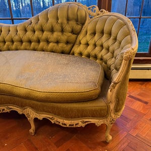 May include: An antique olive green sofa with a tufted back and seat cushions. The sofa has ornate, cream-colored wooden trim and legs. The sofa is placed in front of a large window with a view of trees.