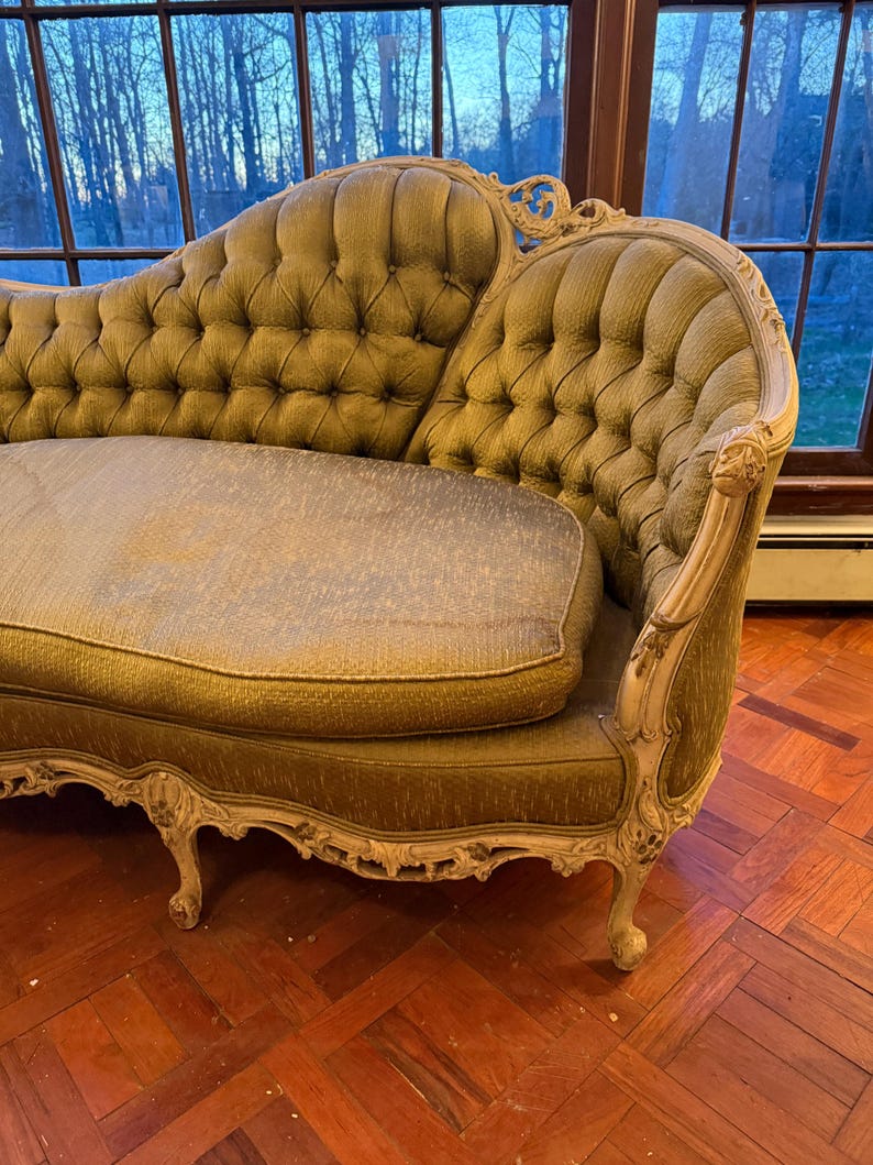 May include: An antique olive green sofa with a tufted back and seat cushions. The sofa has ornate, cream-colored wooden trim and legs. The sofa is placed in front of a large window with a view of trees.