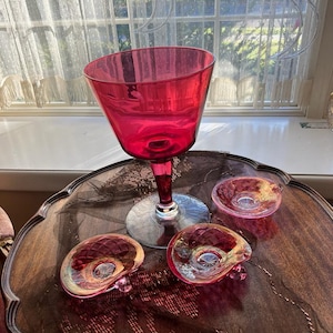 May include: A collection of vintage glassware, including a large, ruby-red stemmed goblet and three smaller, iridescent glass dishes. The goblet has a wide bowl and a clear stem. The dishes are kidney-shaped with a central design.