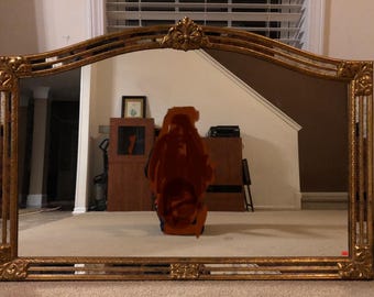 May include: A gold-framed arched mirror with a decorative design. The mirror reflects a room with a wooden cabinet and a person standing in front of it.