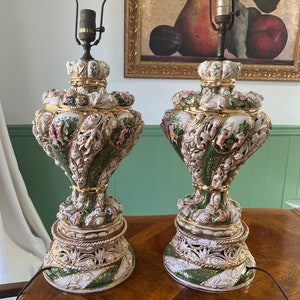 May include: A pair of ornate ceramic table lamps with green, white, and gold accents. The lamps feature intricate details and a floral design.