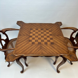 May include: A square wooden game table featuring a central inlaid chessboard pattern with a chevron wood grain border. The table has four extended corner sections with circular indentations and is accompanied by two chairs with curved arms.