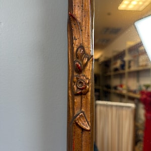 May include: Close-up of a decorative mirror frame. The frame is a warm, metallic gold color with intricate floral and leaf carvings. Red accents highlight the details of the carvings, adding contrast to the design. The mirror reflects a room.