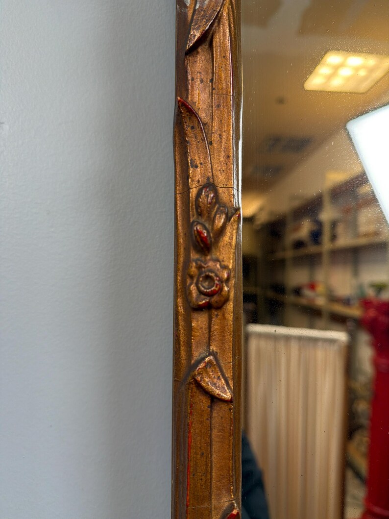 May include: Close-up of a decorative mirror frame. The frame is a warm, metallic gold color with intricate floral and leaf carvings. Red accents highlight the details of the carvings, adding contrast to the design. The mirror reflects a room.