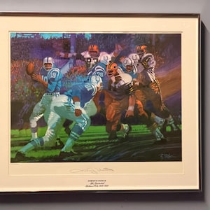 May include: A framed art print depicting a football game with players in blue and white uniforms, including Johnny Unitas. The print features a signature and text reading Johnny Unitas, The Quarterback, Baltimore Colts 1956-1972.