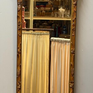 May include: A tall, ornate, rectangular mirror with a gold-colored, carved wooden frame. The top features a decorative bow and a central oval with a floral basket design. The mirror reflects a room with curtains.