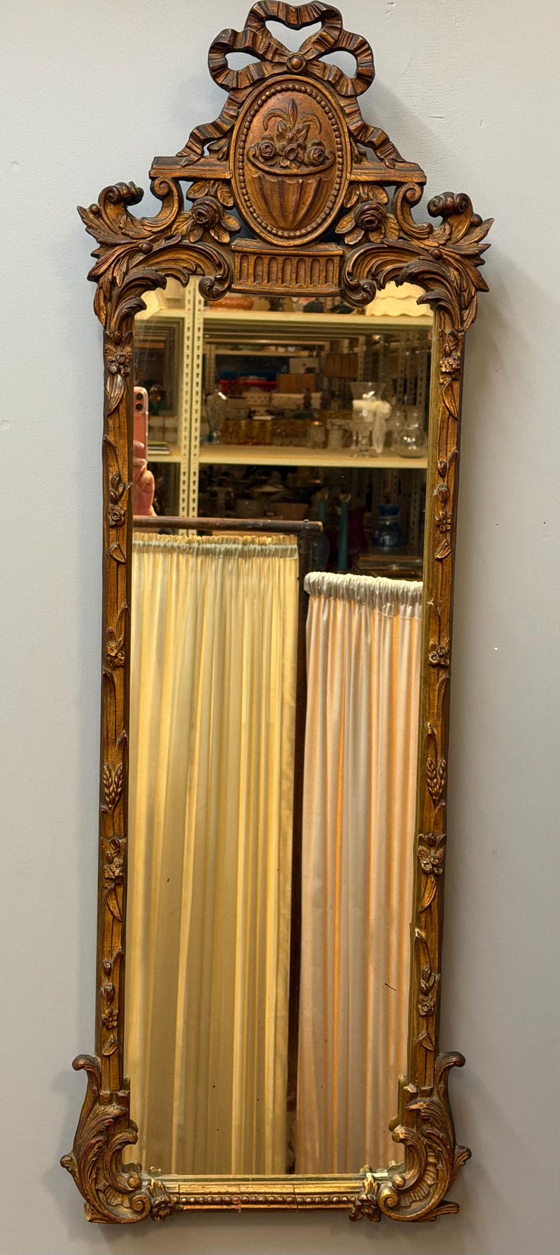 May include: A tall, ornate, rectangular mirror with a gold-colored, carved wooden frame. The top features a decorative bow and a central oval with a floral basket design. The mirror reflects a room with curtains.