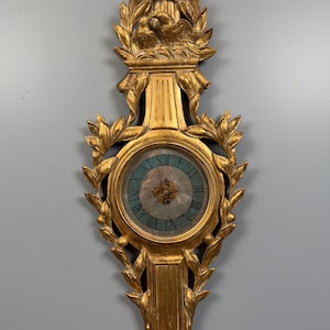 May include: A gold-toned wall clock featuring an ornate, carved frame with leaf motifs and a pair of birds at the top. The clock face has a teal-colored border with black Roman numerals and a central circular design with a gold-colored emblem.
