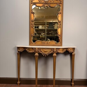 May include: A gold-finished console table with a white marble top and four slender legs, featuring ornate carved details and a central shell motif. Above it hangs a matching rectangular gold-framed mirror with a decorative oval porcelain inlay.
