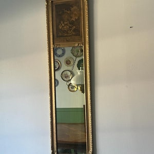 Puede incluir: Un espejo alto de estilo antiguo con marco dorado y un pequeño cuadro enmarcado encima. El espejo refleja una habitación con una mesa de madera y paredes verdes.