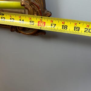 May include: A gold-colored, ornate mirror frame with red accents. A yellow measuring tape shows the frame is approximately 16 inches wide. The mirror is against a gray wall.