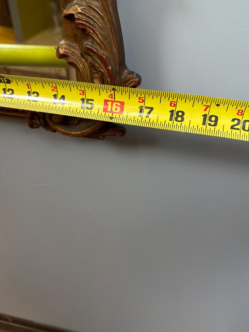 May include: A gold-colored, ornate mirror frame with red accents. A yellow measuring tape shows the frame is approximately 16 inches wide. The mirror is against a gray wall.
