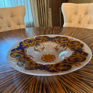 Puede incluir: Un cuenco de vidrio ovalado con interior blanco y borde decorativo. El borde presenta un patrón repetido de diseños dorados y azul oscuro. El cuenco está sobre una mesa de madera marrón oscuro.