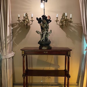 May include: A three-tiered wooden table with gold accents and a decorative lamp with a sculpted base featuring figures and fruit. The lamp is on the top shelf of the table.