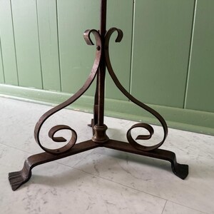 May include: A wrought iron plant stand with a tripod base. The stand features decorative scrollwork and a central vertical support. The metal has a dark, aged finish, and the stand is designed to hold a potted plant.