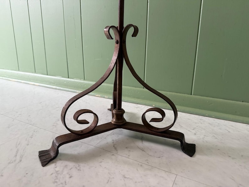 May include: A wrought iron plant stand with a tripod base. The stand features decorative scrollwork and a central vertical support. The metal has a dark, aged finish, and the stand is designed to hold a potted plant.