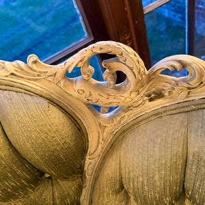 May include: Close-up of a vintage, ornate sofa back. The off-white, carved wooden frame features intricate floral and scroll designs. The seat back is upholstered in a textured, muted green fabric, creating a classic, elegant aesthetic.