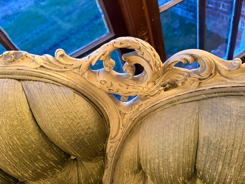 May include: Close-up of a vintage, ornate sofa back. The off-white, carved wooden frame features intricate floral and scroll designs. The seat back is upholstered in a textured, muted green fabric, creating a classic, elegant aesthetic.