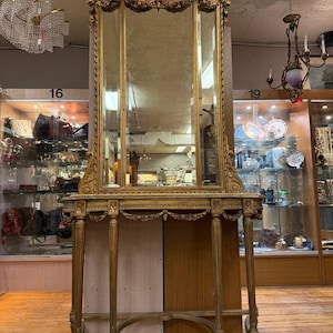 May include: An antique gold-coloured mirror and console table. The mirror has three panels and is topped with an oval painting. The console table has four legs and a decorative apron. The piece is ornate and appears to be vintage.