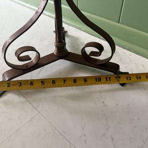 May include: A wrought iron plant stand with a three-legged base and decorative scrollwork. The base measures approximately 15 inches across. The stand has a dark, aged finish and is set on a light-colored floor.