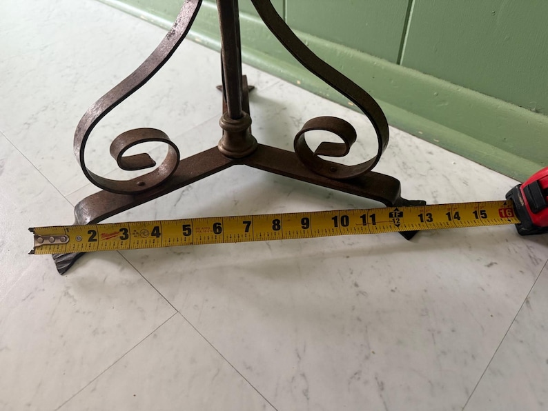 May include: A wrought iron plant stand with a three-legged base and decorative scrollwork. The base measures approximately 15 inches across. The stand has a dark, aged finish and is set on a light-colored floor.