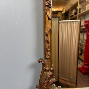 May include: A close-up of a gold-colored mirror frame with ornate carvings and red accents. The frame has a rectangular shape with floral and scroll designs. The mirror reflects a room with shelves and a red pillar.