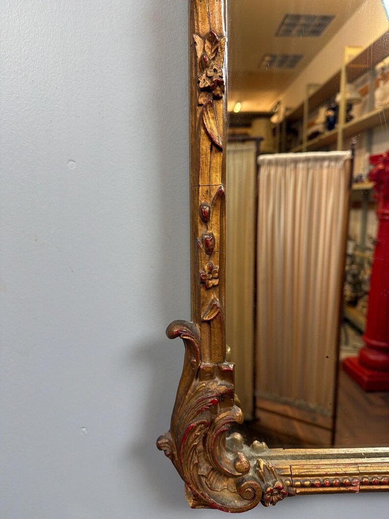 May include: A close-up of a gold-colored mirror frame with ornate carvings and red accents. The frame has a rectangular shape with floral and scroll designs. The mirror reflects a room with shelves and a red pillar.