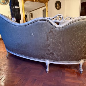 May include: An antique chaise lounge with a curved back and ornate white trim. The upholstery is a muted gray-green color, showing signs of wear. The legs are white and curved, adding to the vintage aesthetic.