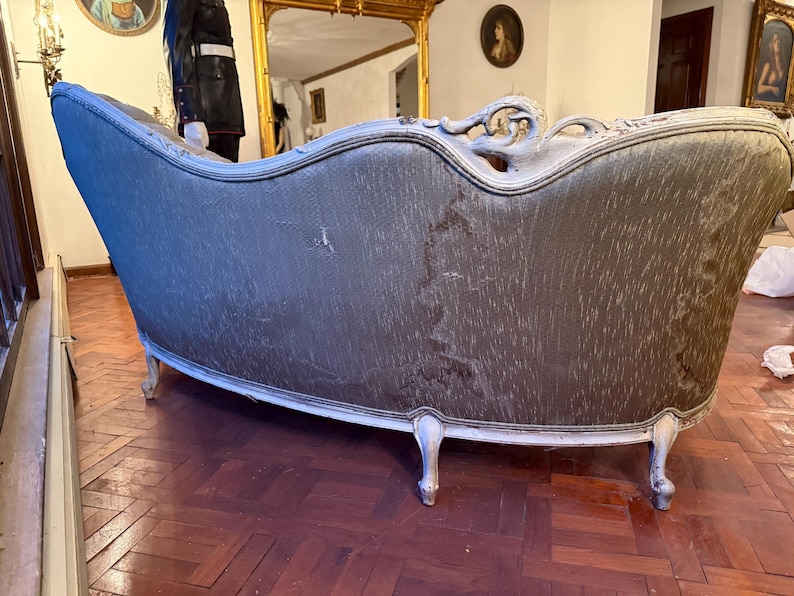 May include: An antique chaise lounge with a curved back and ornate white trim. The upholstery is a muted gray-green color, showing signs of wear. The legs are white and curved, adding to the vintage aesthetic.