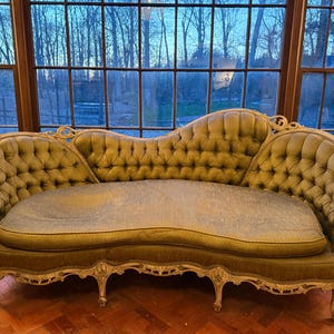 May include: An antique, olive green chaise lounge with a tufted back and seat. The ornate, cream-colored frame features carved details. The sofa is positioned in front of a large window with a view of trees.