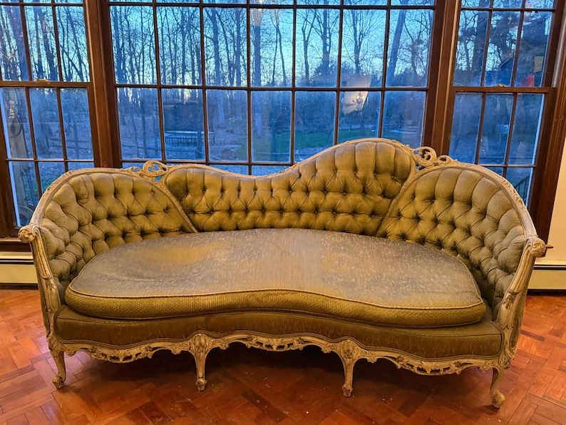 May include: An antique, olive green chaise lounge with a tufted back and seat. The ornate, cream-colored frame features carved details. The sofa is positioned in front of a large window with a view of trees.