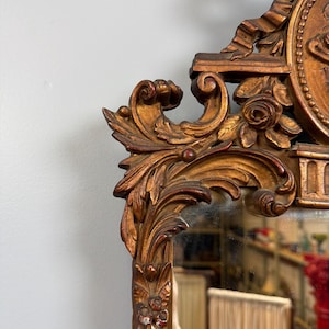 May include: Ornate antique mirror with a detailed, carved wooden frame. The frame is painted in a gold color with red accents, featuring floral and scrollwork designs. The mirror reflects a room with a red column and a tan lampshade.