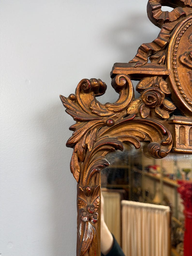 May include: Ornate antique mirror with a detailed, carved wooden frame. The frame is painted in a gold color with red accents, featuring floral and scrollwork designs. The mirror reflects a room with a red column and a tan lampshade.