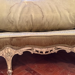 May include: Close-up of a vintage sofa with a distressed, ornate wooden frame. The sofa features a green and gold patterned fabric with visible water stains. The legs and frame are painted in a cream color, showcasing intricate carvings.