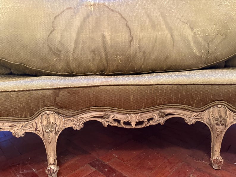 May include: Close-up of a vintage sofa with a distressed, ornate wooden frame. The sofa features a green and gold patterned fabric with visible water stains. The legs and frame are painted in a cream color, showcasing intricate carvings.