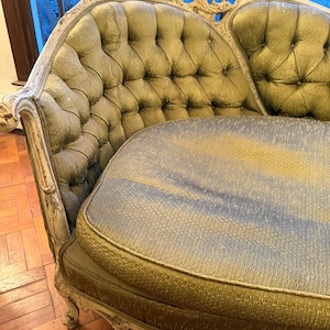 May include: An antique chaise lounge with a tufted back and seat. The upholstery is a muted green with a subtle sheen, and the frame is painted a distressed white. The ornate details and curved design give it a vintage aesthetic.