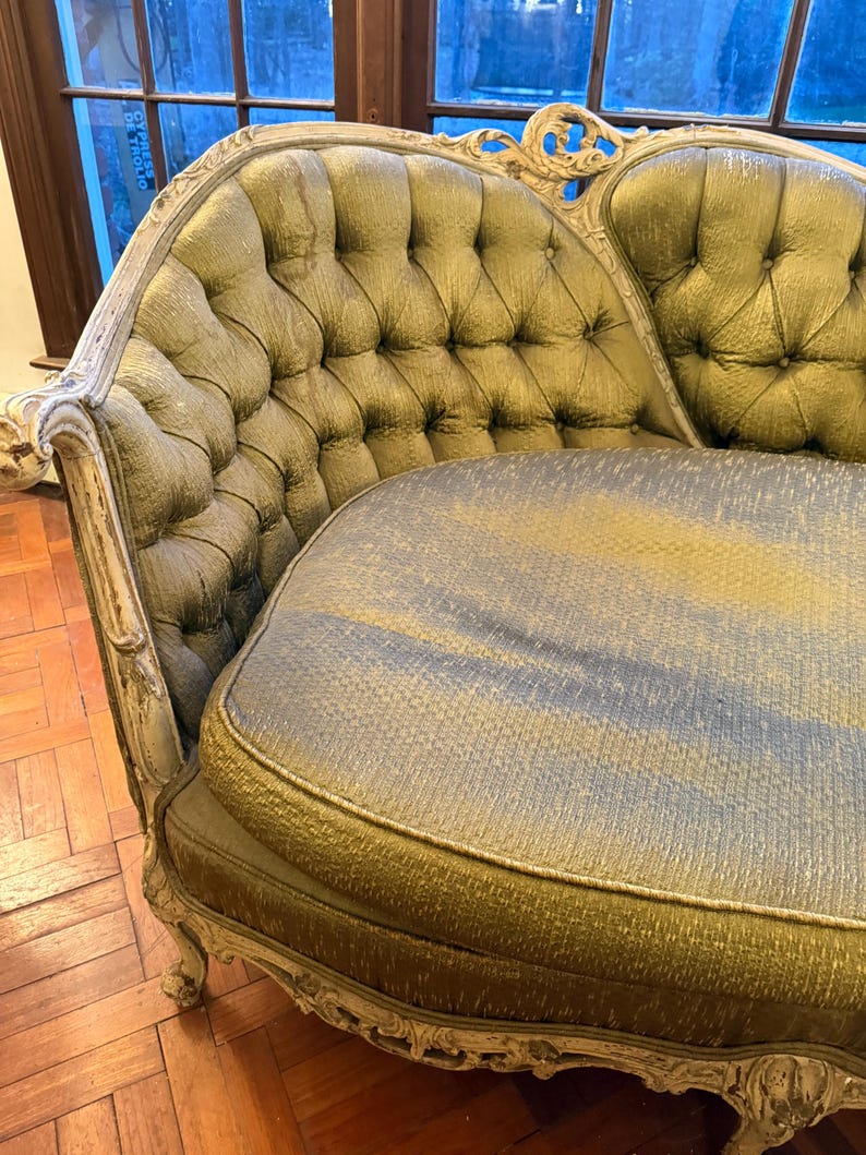 May include: An antique chaise lounge with a tufted back and seat. The upholstery is a muted green with a subtle sheen, and the frame is painted a distressed white. The ornate details and curved design give it a vintage aesthetic.