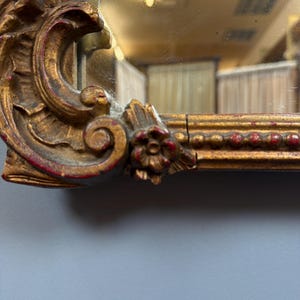 May include: Close-up of a gold-colored mirror frame with ornate, carved details. The frame has a distressed finish with red accents. The mirror reflects a room with a light fixture and curtains. The frame features floral and scroll designs.
