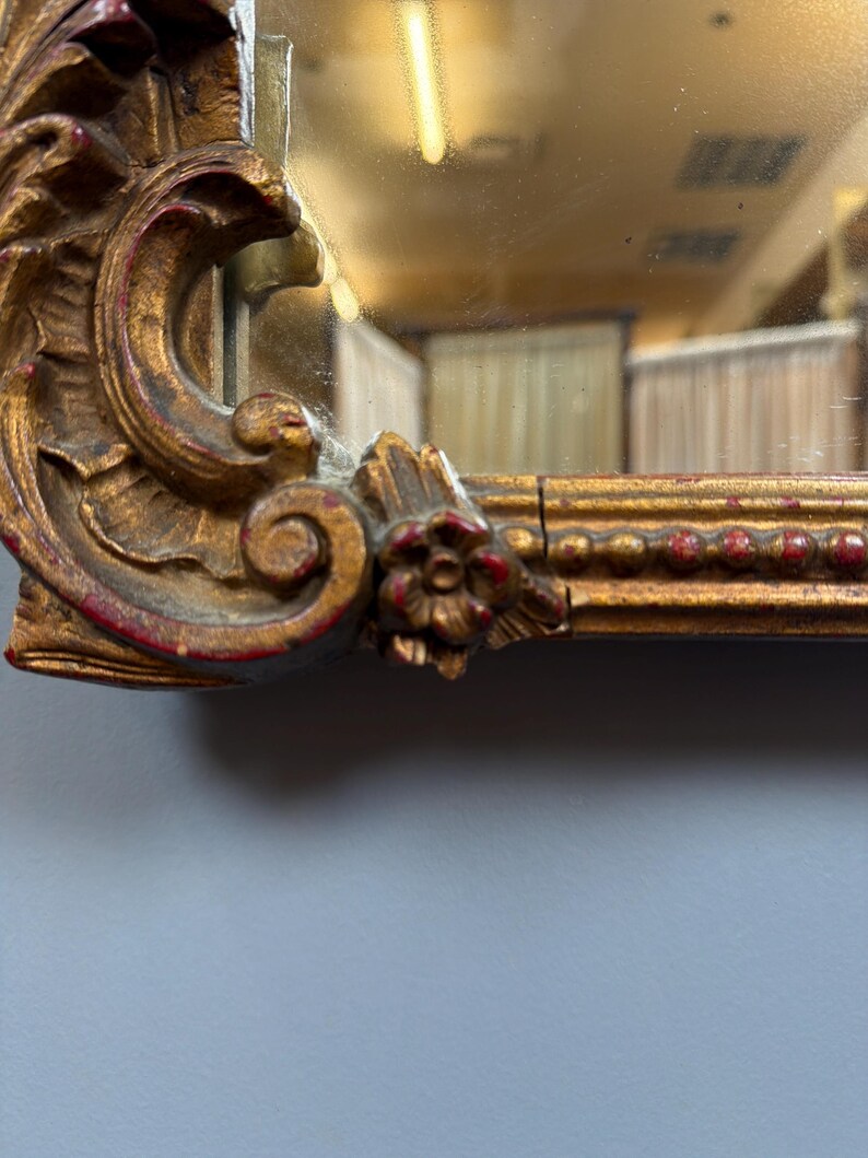 May include: Close-up of a gold-colored mirror frame with ornate, carved details. The frame has a distressed finish with red accents. The mirror reflects a room with a light fixture and curtains. The frame features floral and scroll designs.