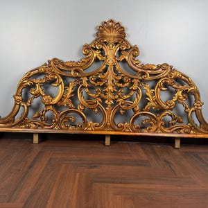 May include: Ornate gold-colored headboard with intricate scrollwork and floral details. The headboard has a curved top with a central shell-like design. The piece is made of wood and has a distressed, antique finish. The headboard is supported by wooden legs.