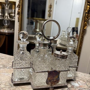 May include: A vintage perfume bottle set featuring five clear glass bottles with crystal stoppers. The bottles are held in a silver-toned metal frame with a handle and a decorative shield. The bottles are made of cut glass.
