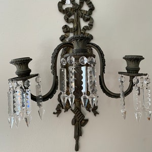 Puede incluir: Una lámpara de pared de dos brazos con un diseño decorativo de lazo en la parte superior. La lámpara tiene prismas de cristal colgando de cada brazo.